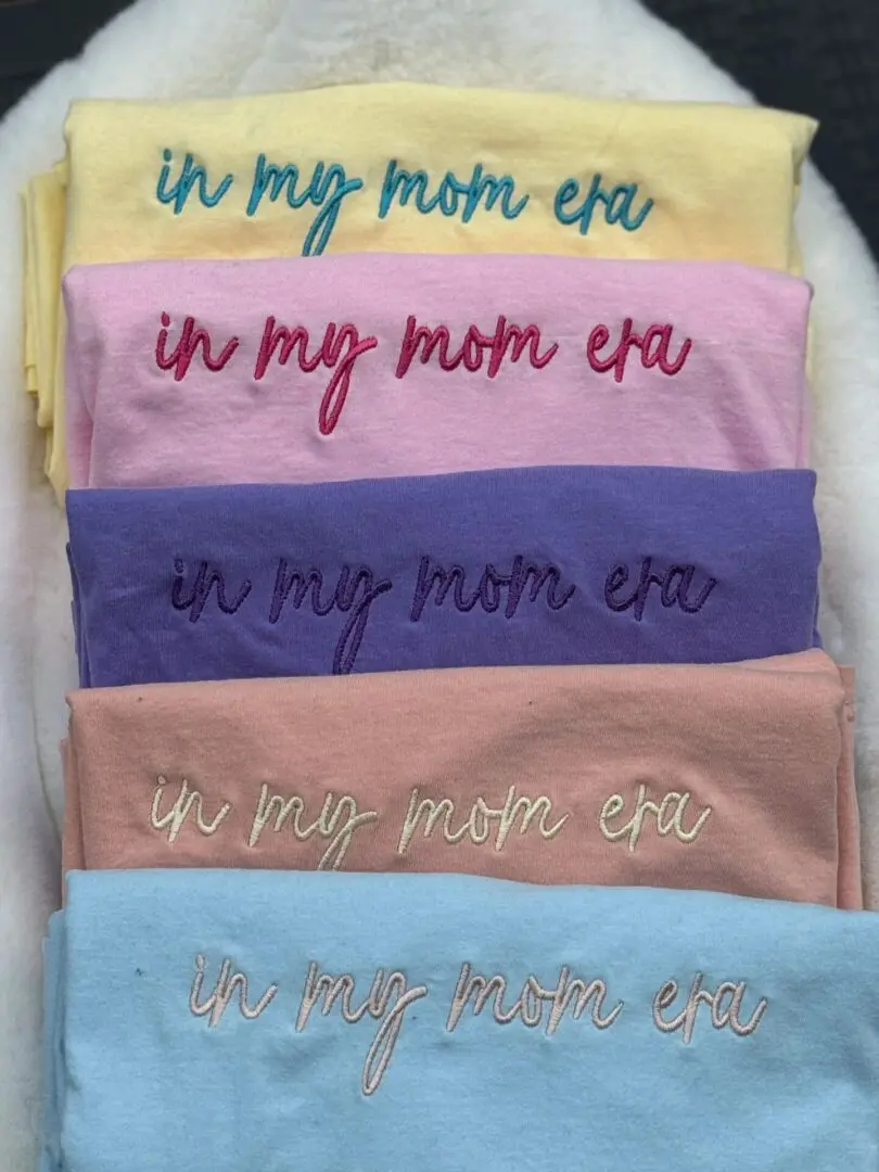 A bunch of different colored shirts with the words in mom era written on them.